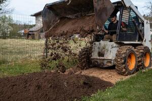 A skid steer loader with orange wheels dumps soil onto the ground, scattering dirt mid air. A wire fence and wooden structures are visible nearby. photo