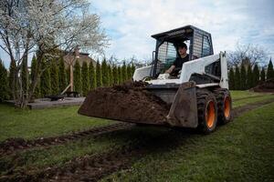 A skid steer loader moves soil on grass, leaving tire tracks. A blossoming tree, trimmed hedges, a wooden platform, and a house roof are visible. photo