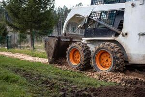 A skid steer loader with orange wheels works on a dirt path, leaving tire tracks. The bucket is lowered, with a wire fence and evergreen trees nearby. photo
