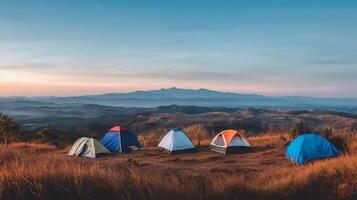 Multiple camping tents are arranged in a scenic mountainous area, surrounded by tall trees and wild grass. photo