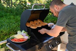 Grilling in the open air a man cooks beef patties and toasts buns next to a plate of crisp vegetables and cheese slices, ready for a summer feast. photo