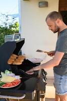 A man grills burger patties over an open flame, with sesame seed buns toasting nearby and a plate of fresh vegetables and cheese ready for burger assembly. photo
