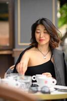 An elegant Asian girl dressed in black relaxes at a terrace cafe table with a cup of coffee. Her serene expression reflects the calm start of the day. photo