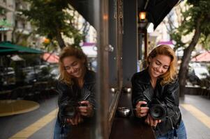 Standing near a cafe, a woman holds her camera while her reflection appears in the glass window. The soft light and double image add depth to the urban atmosphere. photo