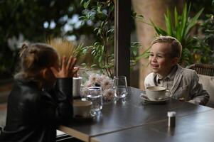 A boy and a girl sit across from each other at a small cafe table, both holding cups of tea, smiling shyly. photo