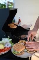 Close-up of hands building burgers by the grill, layering crispy lettuce, tomatoes, onions, and cheese on toasted buns, with fresh toppings within reach photo