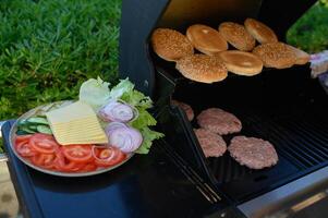 Juicy burger patties and golden buns are grilling outdoors, while a plate of fresh toppings sits ready for burger assembly on a warm summer day photo