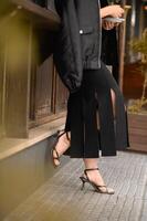 A fashionable woman wearing a summer skirt and heeled sandals stands near a bar counter. Her pose is casual yet poised, capturing a chic, urban vibe in the warm daylight photo