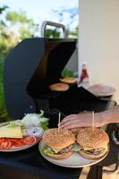 Hands build juicy burgers next to a sizzling grill, stacking fresh lettuce, tomatoes, onions, and cheese, then securing each burger with a wooden stick. photo