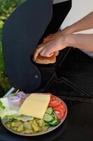 Hands place burger buns on a hot grill next to sizzling patties, with a plate of fresh vegetables and cheese ready for assembling burgers. photo