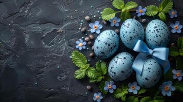 Speckled blue Easter eggs with satin ribbon and small blue flowers arranged with green leaves on dark textured background, creating a fresh spring-themed composition with vibrant holiday contrast photo