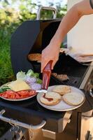 A hand pours ketchup onto toasted burger buns next to a grill where patties are still cooking and a bowl of fresh toppings is ready for assembly. photo