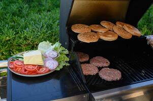 Grill full of sizzling burger patties and toasting sesame buns, with a nearby plate offering crisp lettuce, sliced tomatoes, onions, cheese, and pickles. photo