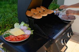 Patio barbecue with sizzling burger patties, golden buns, and a plate of tomatoes, onions, pickles, lettuce, and cheese ready for a summer feast. photo