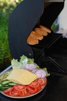 An open grill on a sunny terrace with burger patties and sesame seed buns sizzling, next to a plate of fresh tomatoes, onions, cheese, pickles, and lettuce. photo