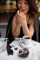 A beautiful woman sits at a cafe table with a cup of coffee, carefully applying red lipstick with a focused expression. photo