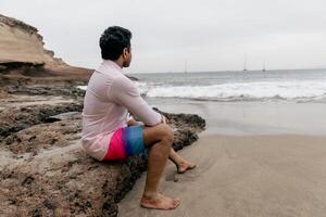 A man sitting on a rock at the beach looking at the ocean and relaxing photo