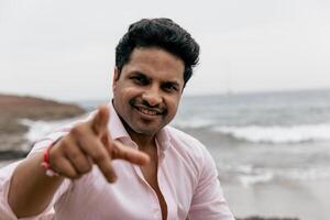 A smiling and confident Indian man pointing towards the camera while at the beach, photo with copy space