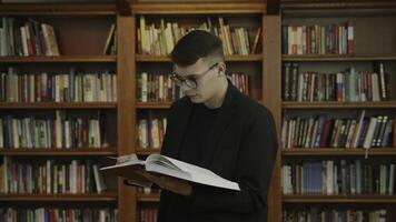 A young man is deeply engrossed in reading a book while seated in a library environment. Media photo