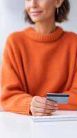Woman in orange sweater holding credit card while sitting at a table, shopping online, enjoying a relaxed moment, financial transaction concept photo