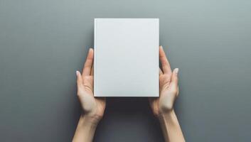 Female hands holding a blank book cover against a gray background, captured from a top view perspective, creating a minimalist mockup photo