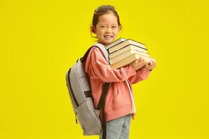 Cheerful little korean first grader girl in casual clothes with many books for studying at school and backpack on shoulders isolated over yellow background photo