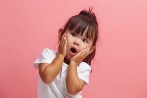 Amazed horrified little three years old girl keeps mouth opened, hands on cheeks, being shocked stares at camera, isolated over pink background. Kid expresses great surprisement. photo