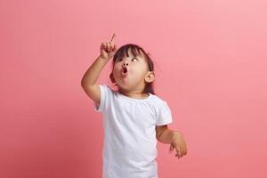 Amazed shocked little girl notices something upwards expresses surprisement looks in amazement points to something up, says wow standing isolated on a pink background photo