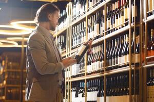 Indoor shot of experienced man sommelier taking wine bottle from shelf, choosing perfect variant for visitor based on his requests. Training for beginners wine experts. photo