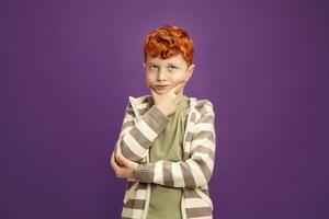 Pensive Red Headed Little Boy Around 7-8 years Puzzled Standing Keeping Hand to Cheek with Thoughtful Face Expression Looking Upwards, Wearing in Casual Clothes Standing on Isolated Violet Background photo
