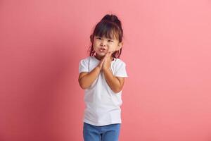 Funny little asian girl keeping palms pressed together in front of her, asks and begging something, has regretful or sorrowful expression standing over pink isolated background photo
