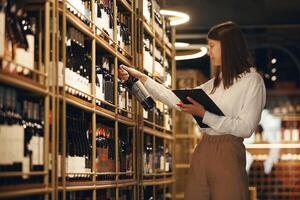 Profile portrait of caucasian young woman sommelier holds bottle of red wine and making notes standing on wine cellar background with various red and white wines on shelf. photo