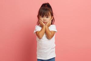 Scared little Asian ethnicity girl looks with fear eyes at camera, being afraid of something, keeps hands on cheeks, looks in terror, isolated over pink background. photo
