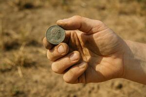 Dusty hand holding ancient green coin over dry soil background. photo