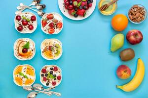 Left 6 yogurts displayed vertically in 2 rows near empty space and around plate of berries, fruits, nuts, honey, 6 spoons on light blue background. Six yogurts, ingredients and empty space. Top view. photo