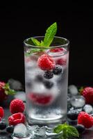 A glass of chilled mineral water with ice cubes and berries near spilled blueberries, strawberries, raspberries and ice cubes on a black background. Mineral water with berries and ice cubes. Vertical. photo