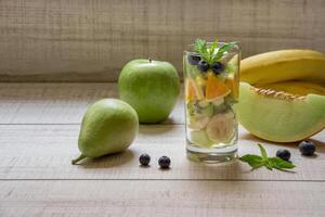 Right glass with sliced fruits melon, banana, apple, kiwi, pear next to ingredients, left empty space for text also in frame blueberries, mint on light background. Glass of fruit salad. Horizontal. photo