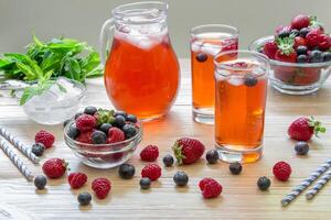 2 compotes of raspberries, strawberries, blueberries, near 2 bowls with berries, bowl of ice, mint leaves, carafe compote, straws on light background scattered berries. Berries compote. Horizontal. photo