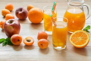 Right fruit juice in glasses with ice near the jar of juice and scattered apricots, peaches, apple, orange left empty space on light wooden background. Apricot peach apple orange juice with ice. photo