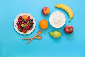 Greek yogurt around fruits orange, banana, pear, peach, apple and a plate with strawberries, raspberries, blueberries and 2 wooden spoons on light blue background. Yogurt, fruits and berries flat lay photo