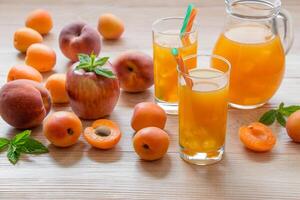 Apricot peach apple juice in glasses with ice near the jar of juice and scattered apricots, peaches, apple, mint on a light wooden background. Apricot peach apple juice with ice. Horizontal. Daylight. photo