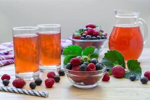 Left two glasses of compote of raspberries, strawberries, blueberries, near bowl with berries, right carafe compote on a light wooden background scattered berries. Fresh berries compote. Horizontal. photo