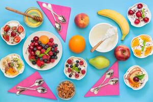6 bowls of yogurt dressed with different ingredients fruit, berries, nuts, honey, and 6 spoons, napkins on light blue background. Six differently dressed yogurts and ingredients. Horizontal. Top view. photo