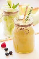 In front a jar with melon smoothie near berries, straws diagonally behind a jar with melon pieces, melon cut slices on a white background. Melon smoothie and salad in 2 jars. Vertical. Close-up. photo