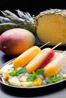 Mango pineapple ice cream popsicles arranged on a decorative plate with crushed ice and fresh fruit. A ripe mango and a halved pineapple enhance the tropical theme. photo