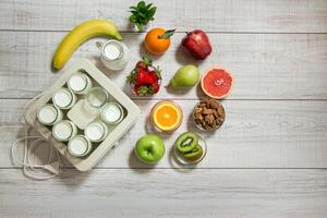 A vibrant collection of fruits including bananas, apples, and berries is placed on a wooden surface alongside a yogurt maker, promoting healthy eating and preparation. photo