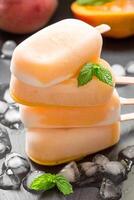 Three mango ice cream popsicles are piled on top of each other on a dark surface. They are surrounded by ice cubes, suggesting a refreshing treat perfect for a warm summer day. photo
