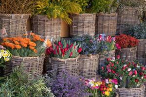 A vibrant assortment of flowers arranged in woven baskets at a market. Brightly colored blooms like tulips, roses, and daisies bring life to the outdoor space, highlighting the beauty of nature. photo