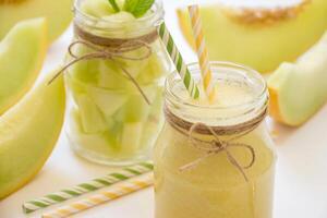 In front a jar with melon smoothie diagonally behind a jar with melon pieces near cut slices of melon, straws on a white background. Melon smoothie and salad in 2 jars. Horizontal. Daylight. Close-up. photo