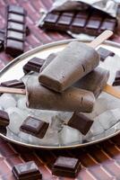 Chocolate ice cream on a stick on a silver platter with ice cubes and pieces of chocolate near pieces of chocolate on a chocolate background. Chocolate ice cream popsicles. Vertical. Close. photo